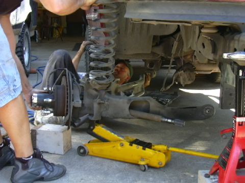 1991 Ford Bronco - Front suspension work in progress on a truck up on jack stands: the wheel is off, exposing the brake rotor, hub and coil spring, while one…