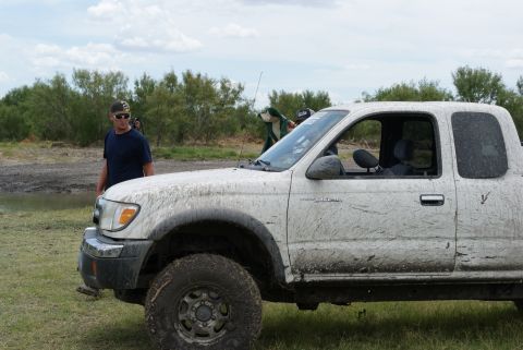 1989 Ford Bronco - A muddy white pickup truck is parked on grassy terrain with trees and a cloudy sky in the background.
