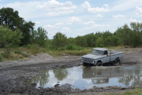 1989 Ford Bronco - A driver navigates a classic pickup truck through a water-filled dirt track under a cloudy sky.