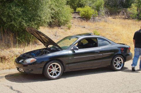 1993 Ford Taurus - A black coupe with its hood open sits on a roadside, showing off its engine and aftermarket wheels.