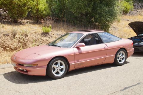 1993 Ford Taurus - A pink Ford Probe GT is parked on the side of a road with trees and a hillside behind it.