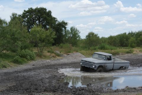 1989 Ford Bronco - A vintage gray pickup truck navigates a muddy terrain, its grille suggesting a 1960s model year.