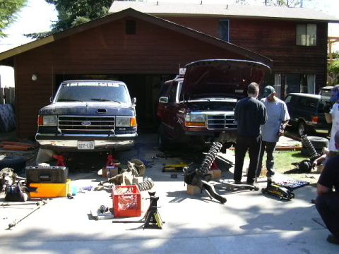1991 Ford Bronco - Driveway suspension work on two Ford trucks parked side by side in front of a brown wood-sided house, one dark blue/grey with a Colorado pl…
