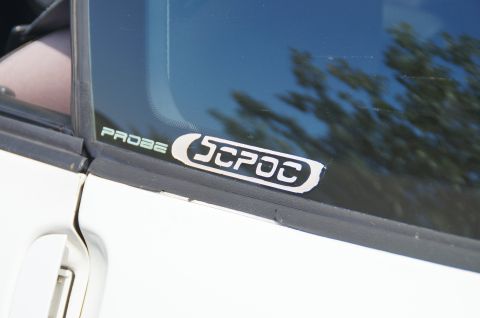 1993 Ford Taurus - A white car door and window display "PROBE" and "SPOC" stickers, with reflections of trees in the glass.