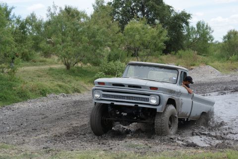 1989 Ford Bronco - A person drives a gray pickup truck through a muddy area, kicking up water and mud.