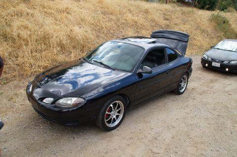 1993 Ford Taurus - A black coupe with its trunk open is parked on a dirt road, with another dark vehicle visible behind it.