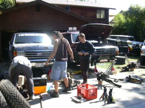 1991 Ford Bronco - Driveway suspension work in progress on two full-size Ford trucks parked outside a brown wood-sided house, with three people working around…