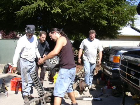1991 Ford Bronco - Driveway suspension swap in progress: four people work around a removed coil-spring front axle assembly with brake rotors and hubs laid out…