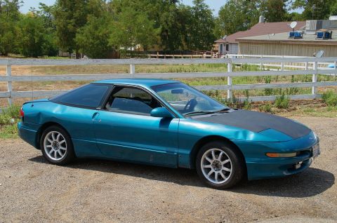 1993 Ford Taurus - Teal coupe with black hood parked on gravel in front of a white fence and buildings.