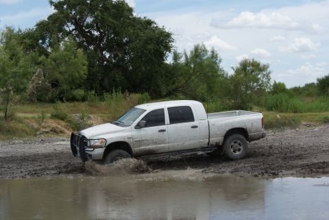 1989 Ford Bronco - A white pickup truck navigates through a muddy off-road area, kicking up water.