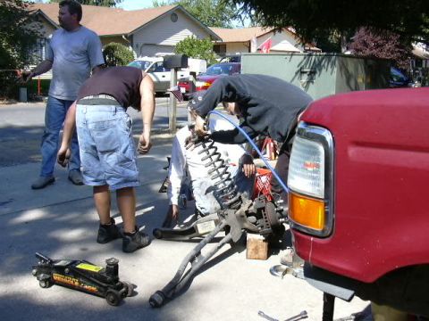 1991 Ford Bronco - Three people work on a suspension swap in a suburban driveway, with a solid front axle, radius arms and tall coil springs laid out on the p…