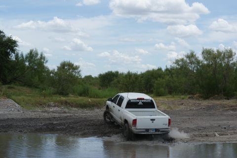 1989 Ford Bronco - A white pickup truck navigates a muddy off-road course under a cloudy sky.