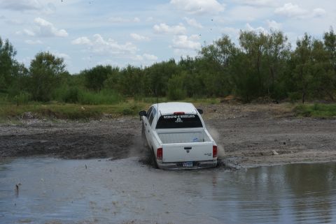 1989 Ford Bronco - A white pickup truck navigates through a muddy water obstacle, kicking up spray from its tires.