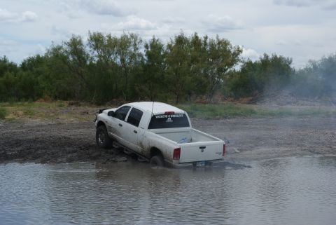 1989 Ford Bronco - A white pickup truck is stuck in a muddy body of water with trees in the background.