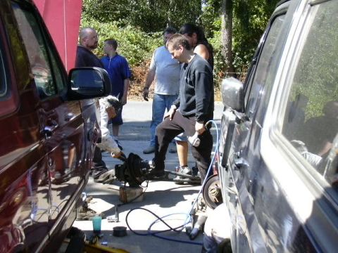 1991 Ford Bronco - Driveway wrenching session between two trucks with their front wheels removed, a dark red one on the left with its tow mirror in frame, and…