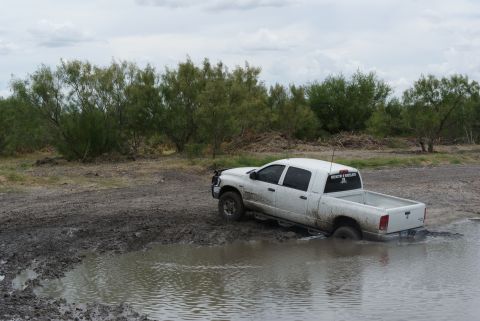 1989 Ford Bronco - A white pickup truck is stuck in a muddy area with water, surrounded by vegetation under a cloudy sky.
