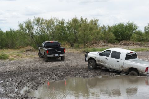 1989 Ford Bronco - Two pickup trucks are stuck in a muddy area, with one partially submerged in a water-filled rut.