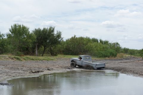 1989 Ford Bronco - A gray pickup truck navigates through a muddy, watery terrain under a cloudy sky.