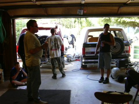 1991 Ford Bronco - View out of an open garage door where several people stand around during a work session, with the rear of a two-tone dark and white Ford Br…