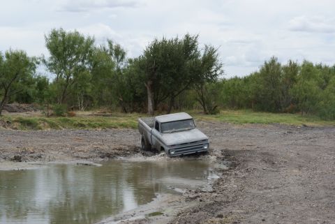 1989 Ford Bronco - A classic gray pickup truck is stuck in a muddy puddle, surrounded by brush and trees under a cloudy sky.