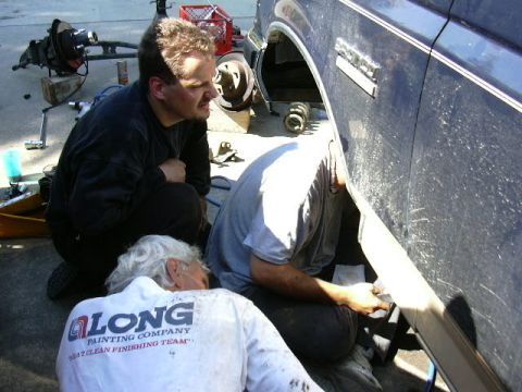 1991 Ford Bronco - Three men crouch beside a dark blue truck with its front wheel removed, working inside the open wheel well where a coil spring and radius a…