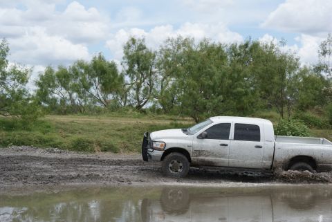 1989 Ford Bronco - A white pickup truck splashes through a muddy area with trees and a cloudy sky overhead.