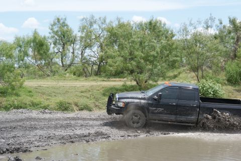 1989 Ford Bronco - A black pickup truck navigates a muddy terrain, kicking up water and mud as it drives through a puddle.