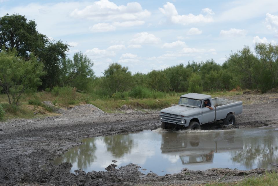 Gray pickup truck driving through mud puddle, driver visible, trees and cloudy sky in background