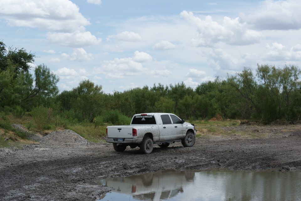 White pickup truck with crew cab and long bed driving on a muddy, wet road with trees in the background. Text on rear window
