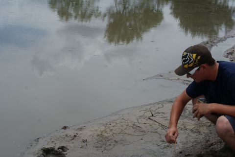 1989 Ford Bronco - A person kneels by the edge of a body of water, observing something on the muddy shore.