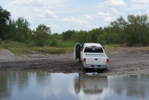 1989 Ford Bronco - A white pickup truck is partially submerged in muddy water with its driver's door open.