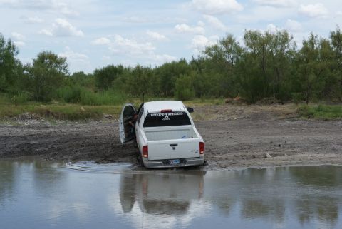 1989 Ford Bronco - A white pickup truck is stuck in a muddy puddle with its driver's door open.