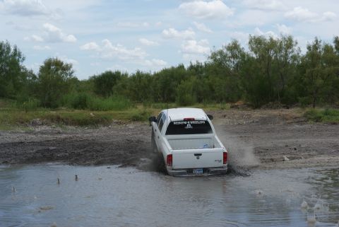 1989 Ford Bronco - Pickup truck navigates through a muddy off-road area under a cloudy sky.