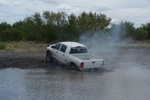 1989 Ford Bronco - A white pickup truck is stuck in a muddy puddle, with smoke billowing from its rear.