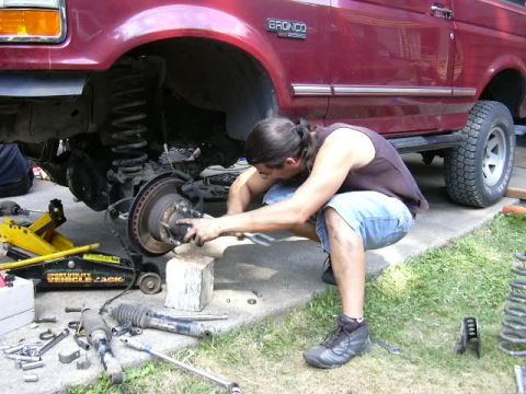 1991 Ford Bronco - A person kneels on a concrete driveway working on the exposed front hub and brake rotor of a maroon Ford Bronco, with the front wheel remov…