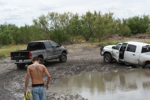 1989 Ford Bronco - Two pickup trucks are stuck in the mud, with a man walking away from one of them in a rural outdoor setting.