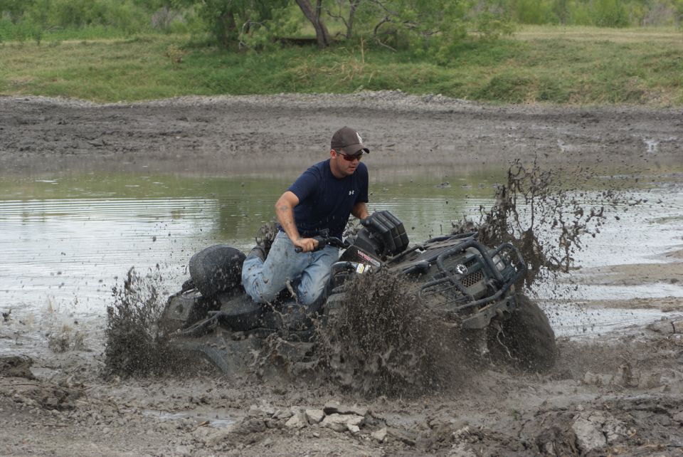 Man riding an ATV through a muddy puddle, kicking up a lot of spray