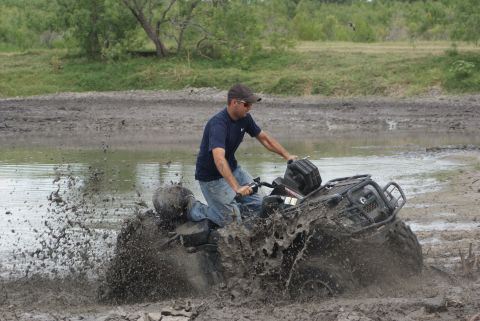 1989 Ford Bronco - A man rides an ATV through a muddy field, kicking up a large spray of water and mud.