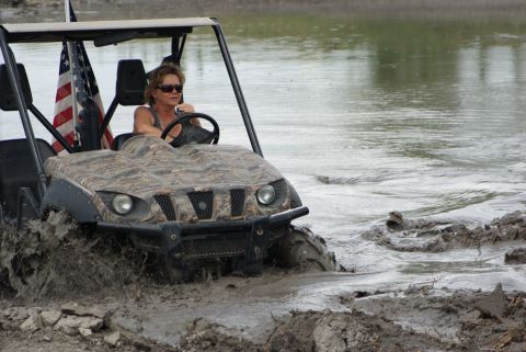1989 Ford Bronco - Woman drives a camouflage UTV through a muddy water obstacle with an American flag attached.