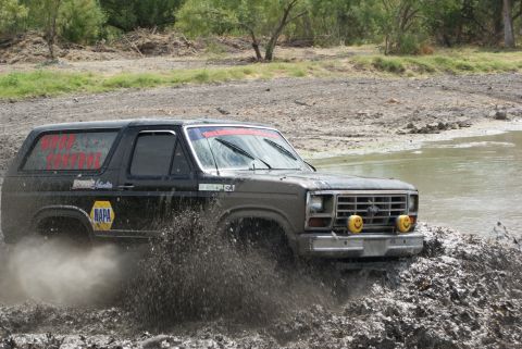 1989 Ford Bronco - Black Ford Bronco navigates a muddy terrain, displaying NAPA branding and red text on its windows.