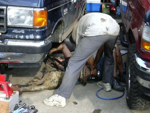 1991 Ford Bronco - Two individuals are actively working on the suspension of a blue truck, with a red truck also visible in the scene.
