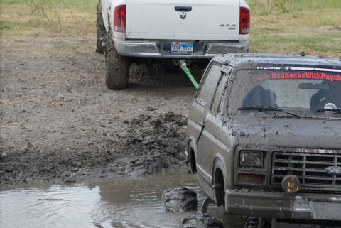 1989 Ford Bronco - A muddy SUV is stuck in a puddle while a white pickup truck tows it out.