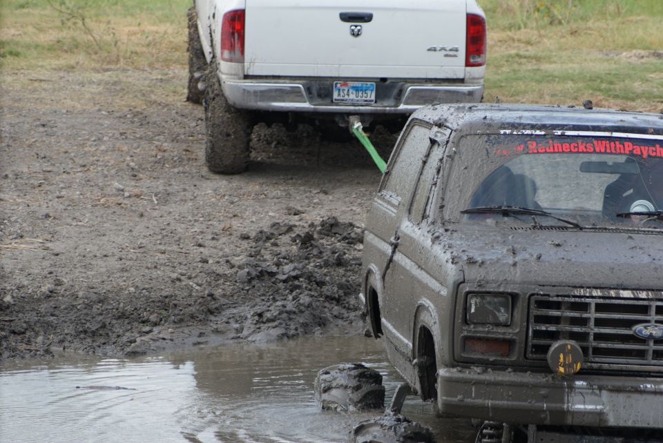 Mud-covered SUV with a red sticker on the windshield, stuck in mud and being towed by a white pickup truck