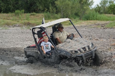 1989 Ford Bronco - Two people ride in a muddy side-by-side utility vehicle as it navigates through a muddy terrain.