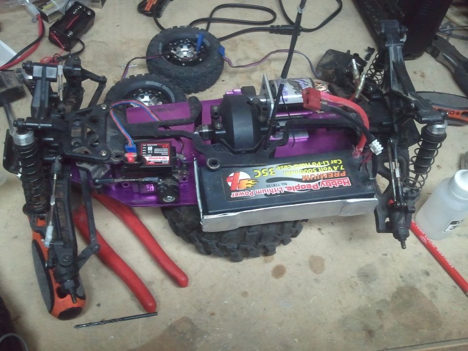 RC car chassis with purple frame, battery pack, and suspension components. Tools and drill bit are on the workbench
