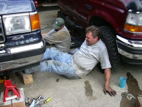 1991 Ford Bronco - Two mechanics work on a dark blue Ford F-series truck, which appears to be a 1992 model, not a 1991 Bronco.
