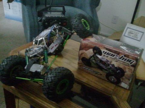 1985 Ford Bronco - A remote control rock crawler with green suspension components sits on a wooden surface next to its product box, with another RC vehicle in…