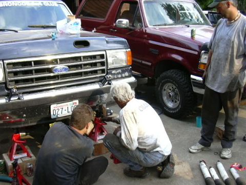 1991 Ford Bronco - Two men work on the front suspension of a dark-colored Ford truck, with a red Ford Bronco parked behind it.
