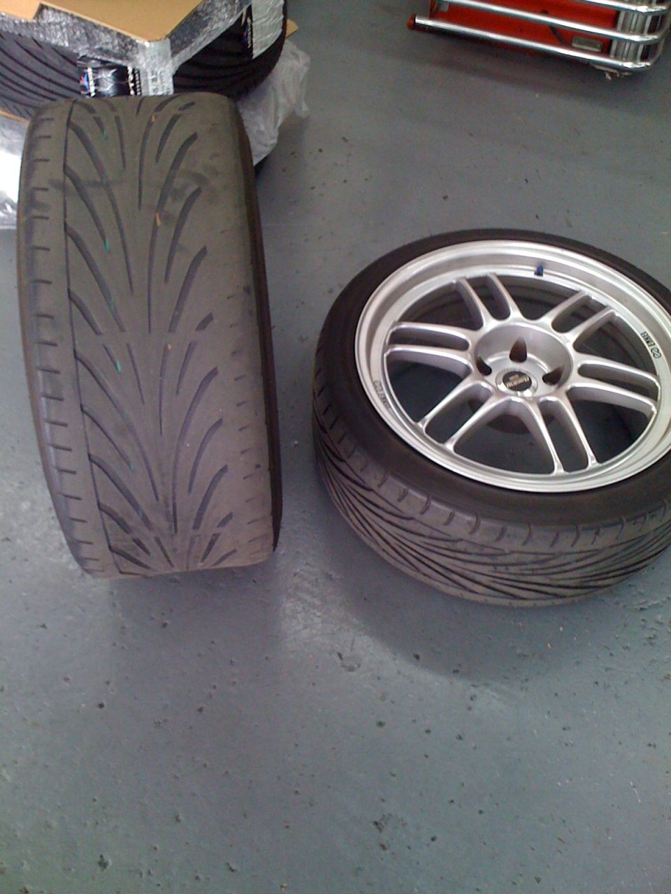 One tire is on its side, the other is mounted on a silver 5-spoke wheel. Text on the wheel reads 'ENKEI'