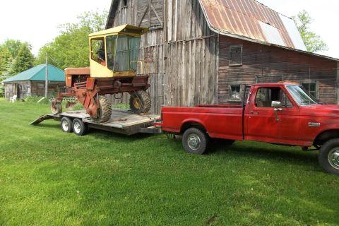 1987 Ford F-250 - A red 1987 Ford F-350 pickup truck is seen from the side, towing a trailer with a piece of farm machinery on it in front of a barn.